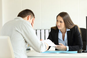 Two people sitting at an office desk. One person has his head in his hand with the other person looking.